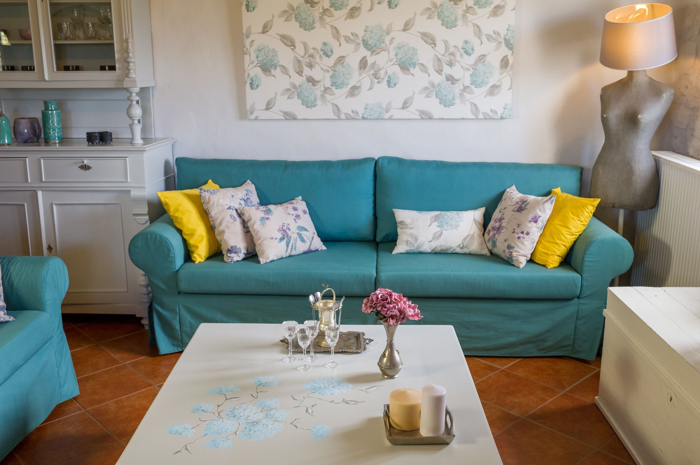 Ein blaugrünes Sofa mit floralen Kissen, ein weißer Couchtisch mit floralem Muster und eine Vase mit rosa Blumen.