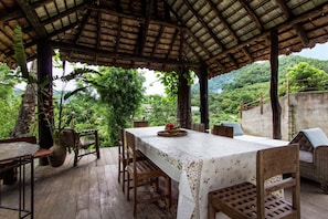 Un área de estar al aire libre techada con una mesa y sillas, rodeada de exuberante vegetación y un paisaje montañoso.