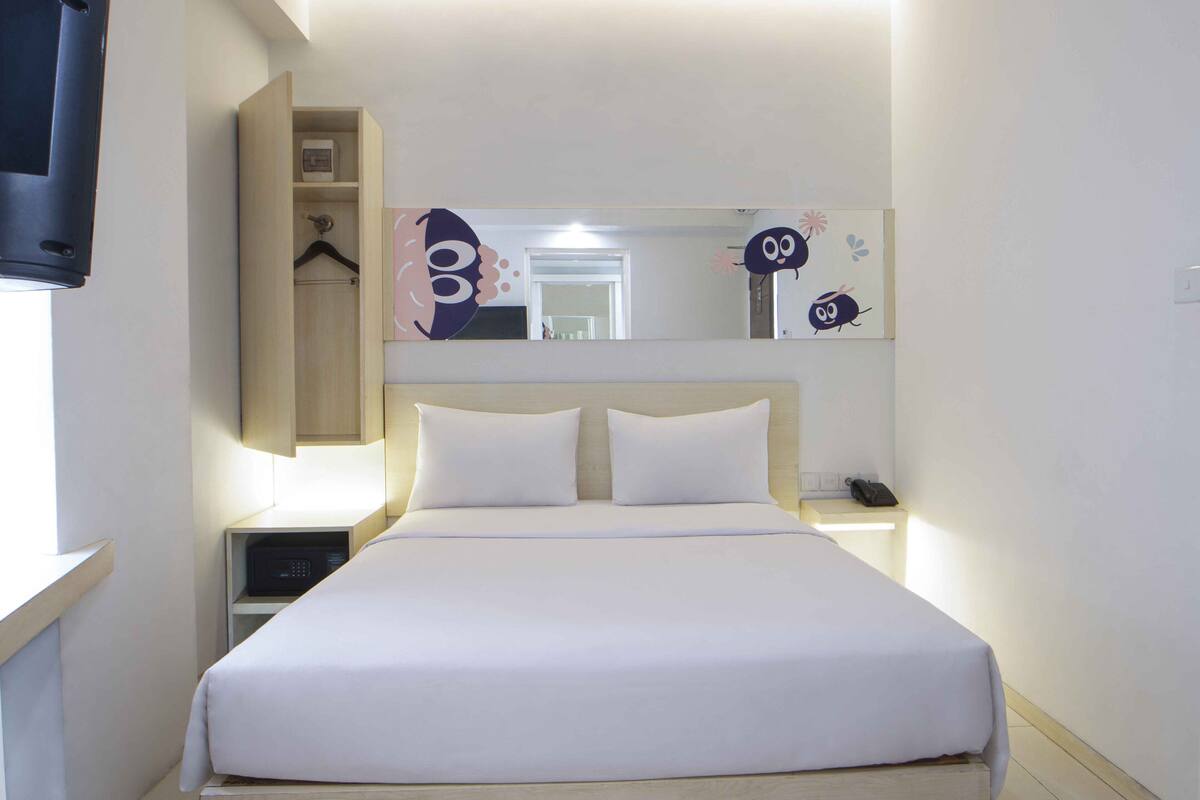 Una cama bien hecha con sábanas blancas, una mesita de noche con lámpara, un estante con un reloj, y un espejo sobre la cama.
