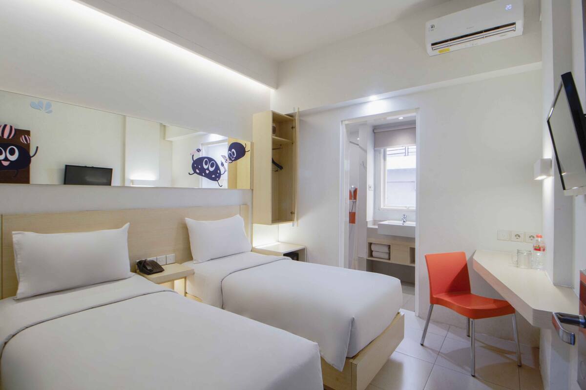 Habitación de hotel con dos camas, un escritorio, una silla, televisión y baño visible a través de una puerta abierta.