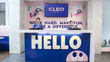 A hotel reception desk with a 'HELLO' sign, a television screen above displaying 'CLEO HOTELS', and a wall with a motivational message.