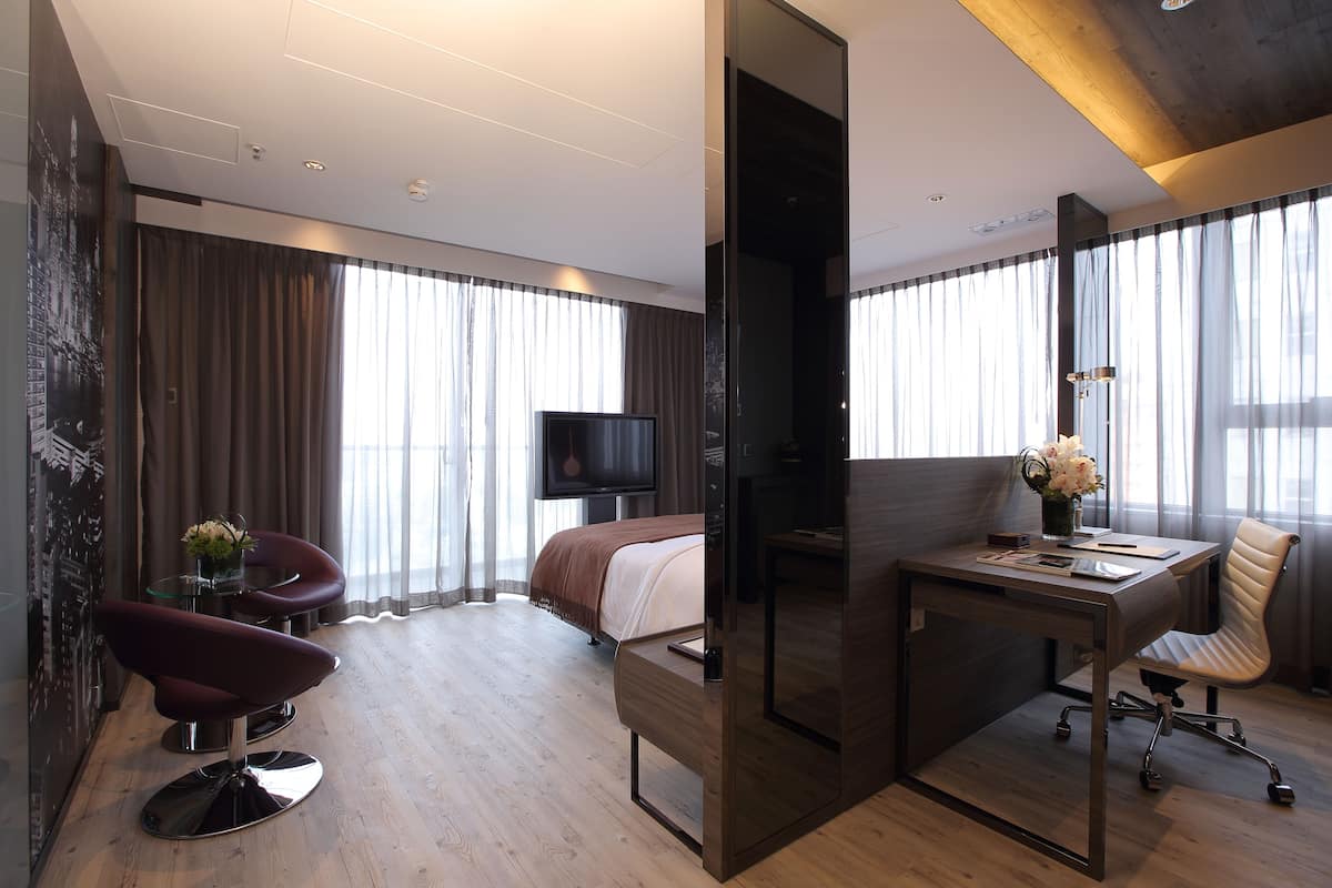A modern hotel room with a wooden floor, a bed with a brown headboard, a desk with a chair, a TV, and large windows with curtains.