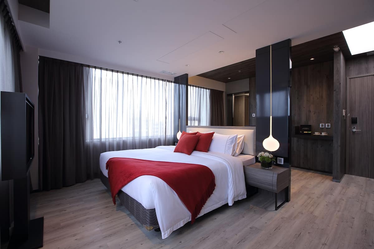 A modern hotel room with a large bed, a bedside table with a lamp, a TV, and a view of the city through large windows.