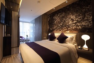 A modern hotel room with a large bed, a mounted TV, a desk with a lamp, and a wall with a tree mural.