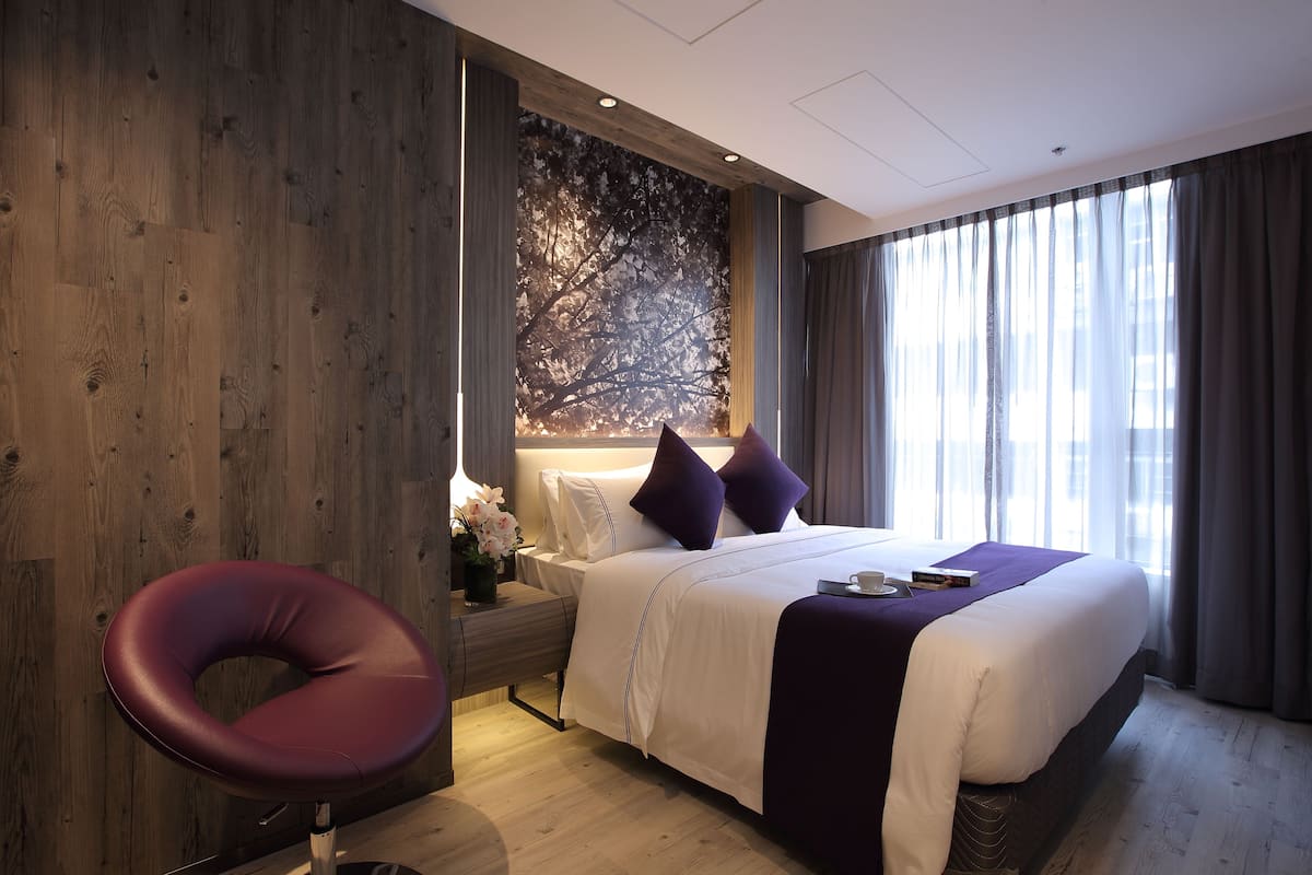 A modern hotel room with a large bed, a wooden accent wall, a dark-colored chair, and a large window with curtains.