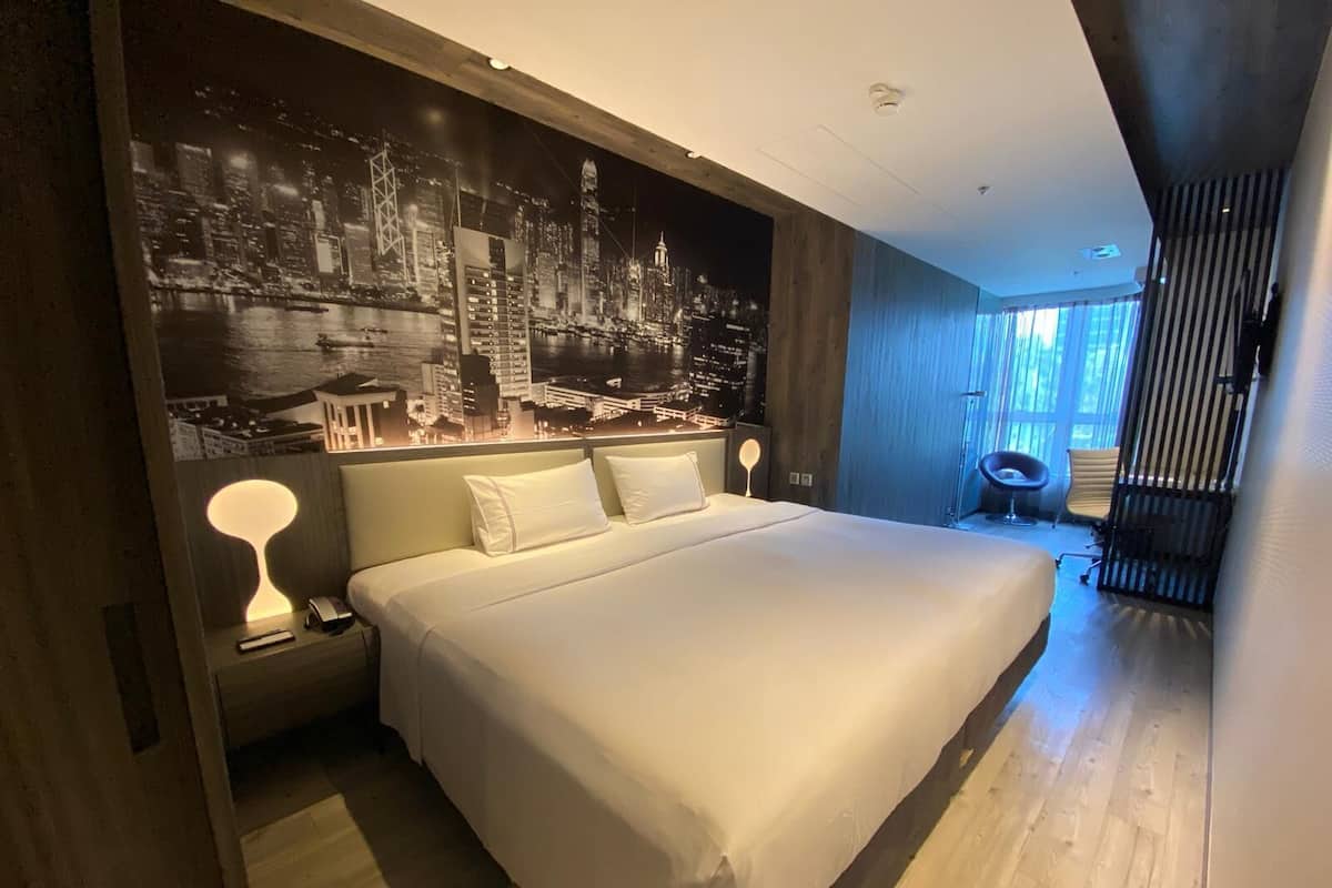 A modern hotel room with a large bed, a cityscape mural on the wall, and a view of the outdoors.