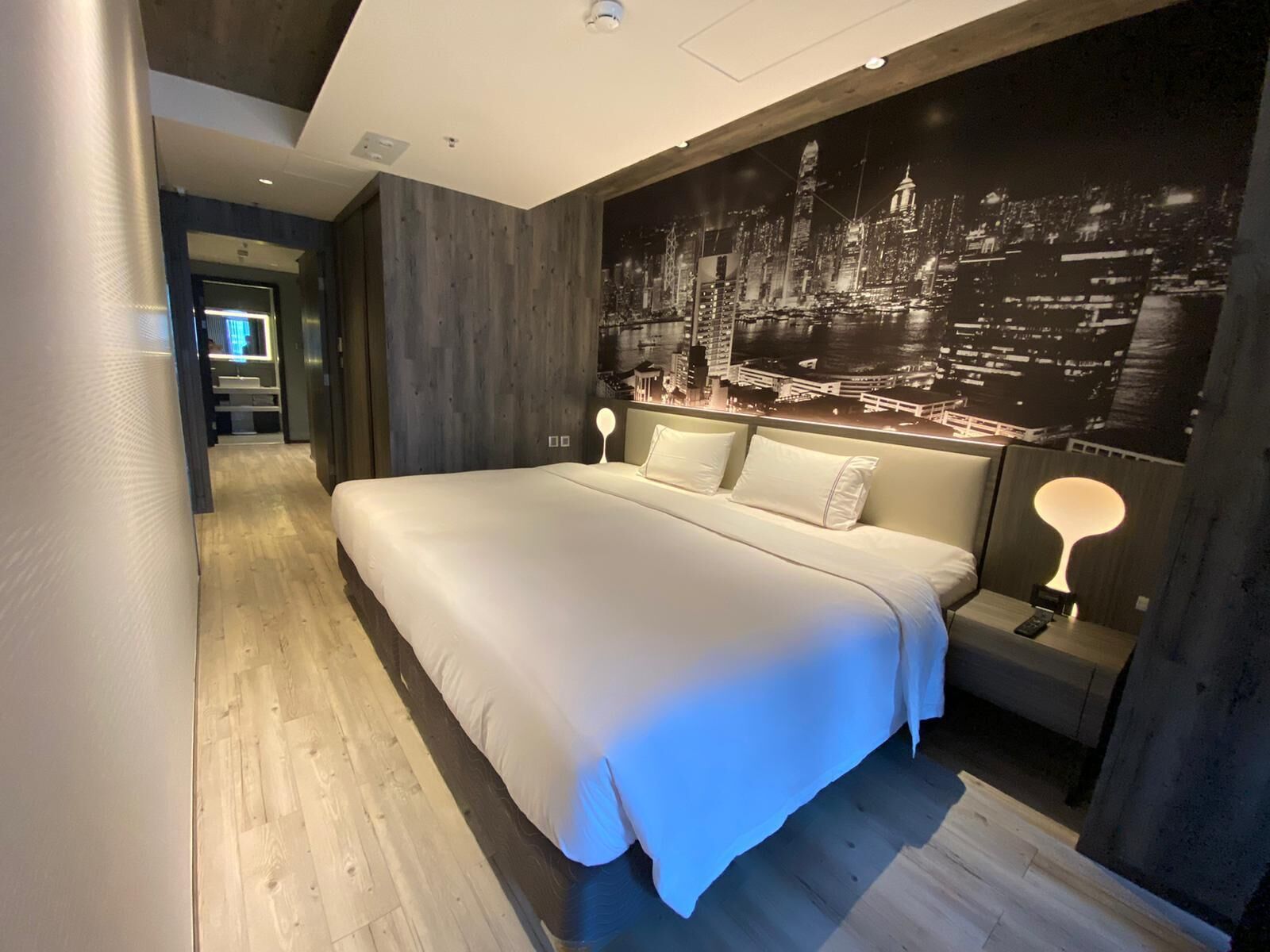Habitació d'hotel moderna amb un llit gran, un mural de paisatge urbà a la paret i un sòl de fusta.