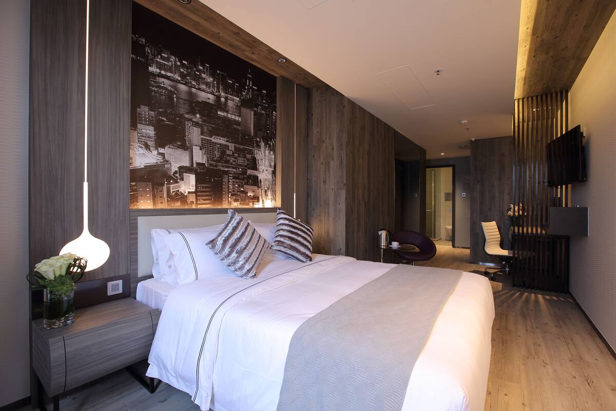A modern hotel room with a large bed, a nightstand with a vase of flowers, a wall-mounted TV, and a cityscape mural.