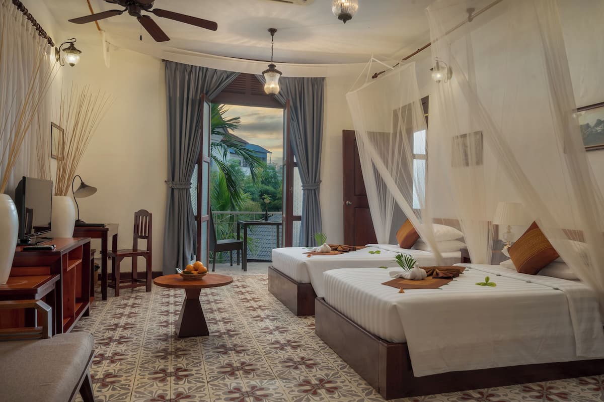 Une chambre avec deux lits, un ventilateur de plafond, une table avec des fruits, une chaise et une vue sur la verdure à l’extérieur.
