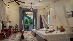 A room with two beds, a ceiling fan, a table with fruits, a chair, and a view of greenery outside. - La Rivière d' Angkor Resort (Siem Reap)