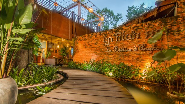 A wooden pathway leading to a brick wall with a sign, surrounded by lush greenery and a small pond. - La Rivière d' Angkor Resort (Siem Reap)