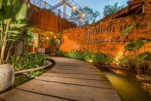A wooden pathway leading to a brick wall with a sign, surrounded by lush greenery and a small pond.