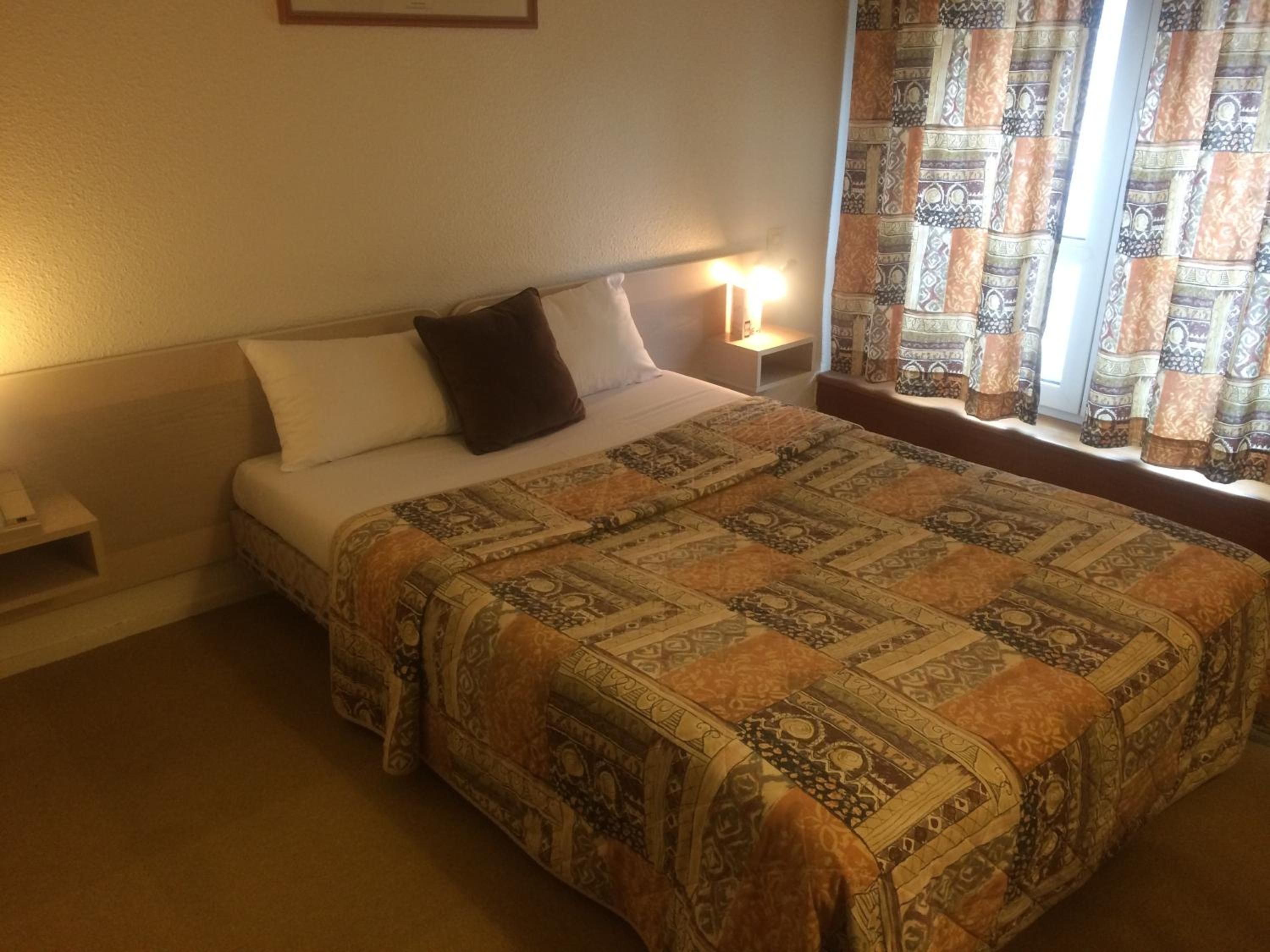Quarto de hotel com cama, mesa de cabeceira, abajur e cortinas estampadas.