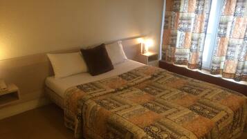 Quarto de hotel com cama, mesa de cabeceira, abajur e cortinas estampadas.