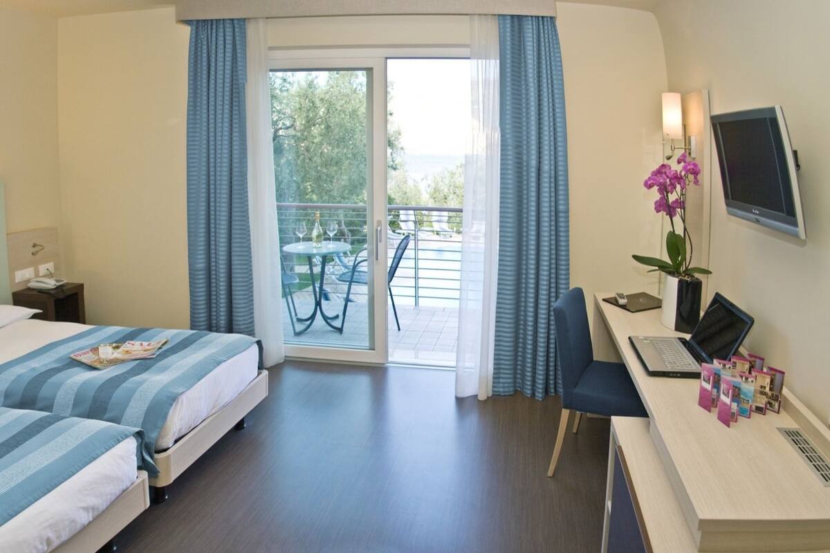 A hotel room with two beds, a desk with a laptop, a TV, and a balcony with a table and chairs.
