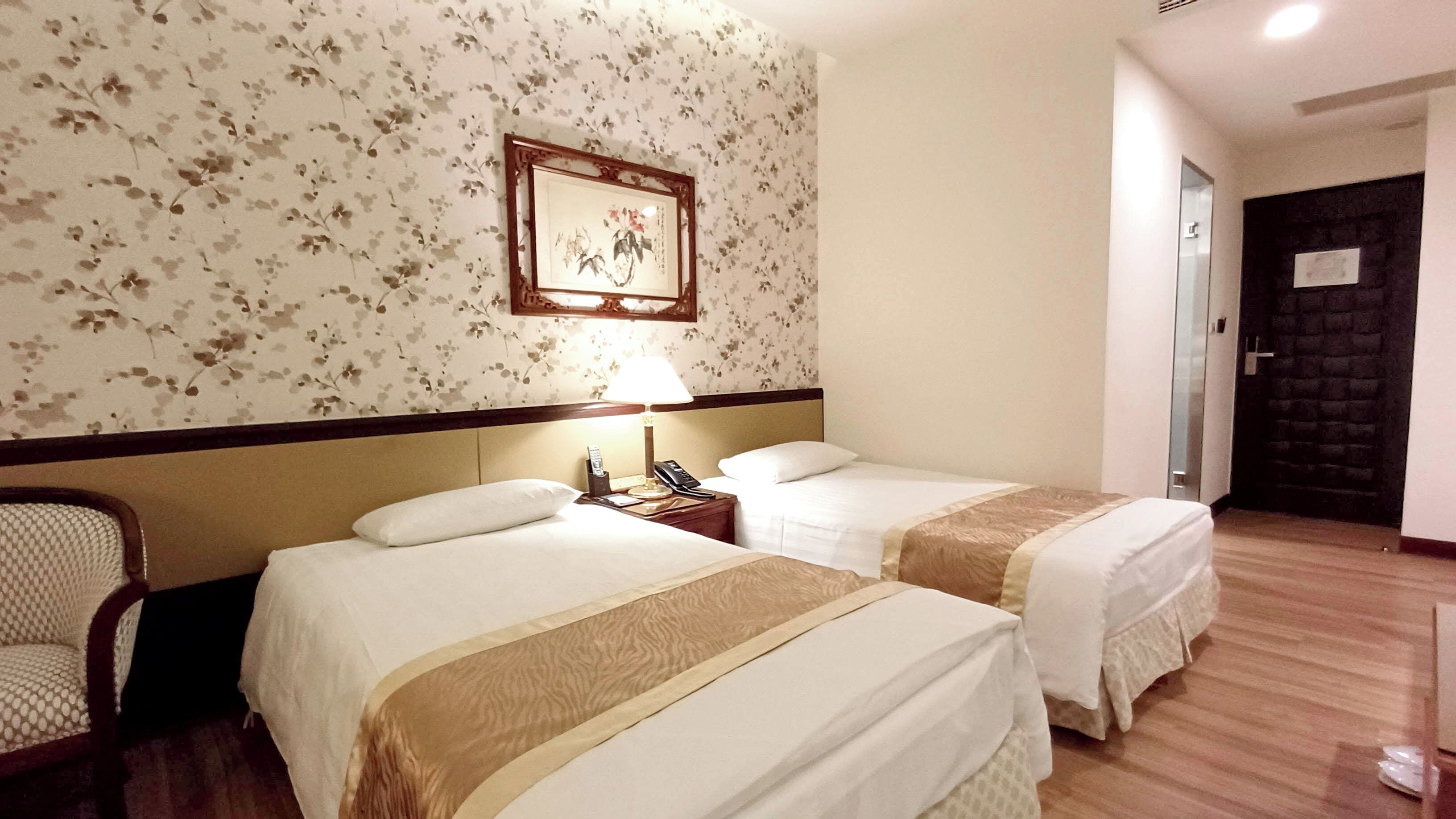 A hotel room with two beds, a nightstand with a lamp, a chair, and a floral wallpaper.