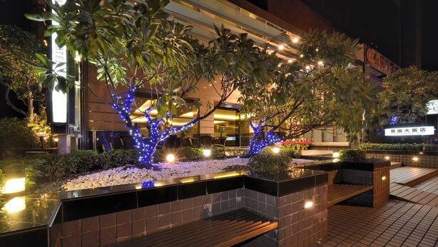 Un espacio exterior bien iluminado con un banco, luces decorativas y un edificio al fondo.