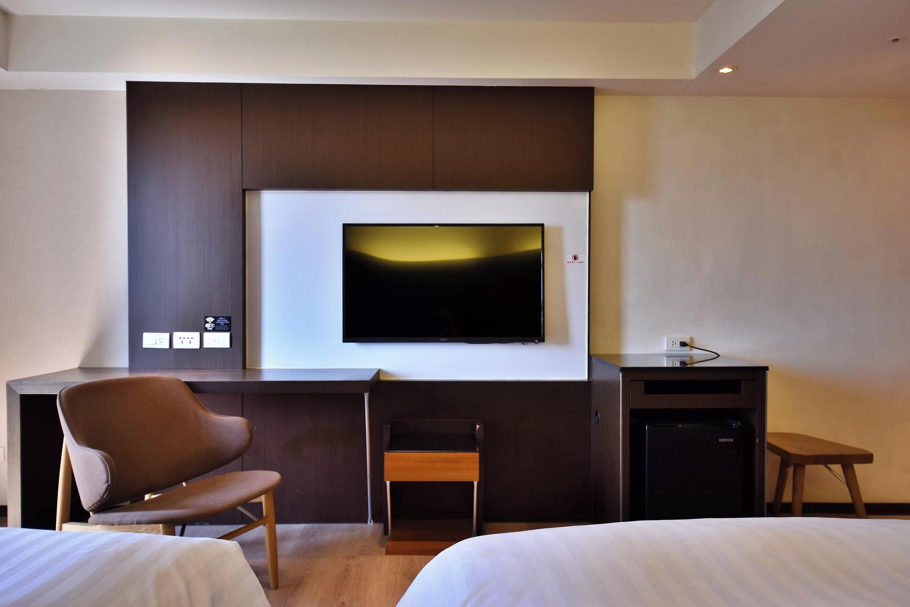 A hotel room with a bed, a desk with a chair, a television, and a small refrigerator.