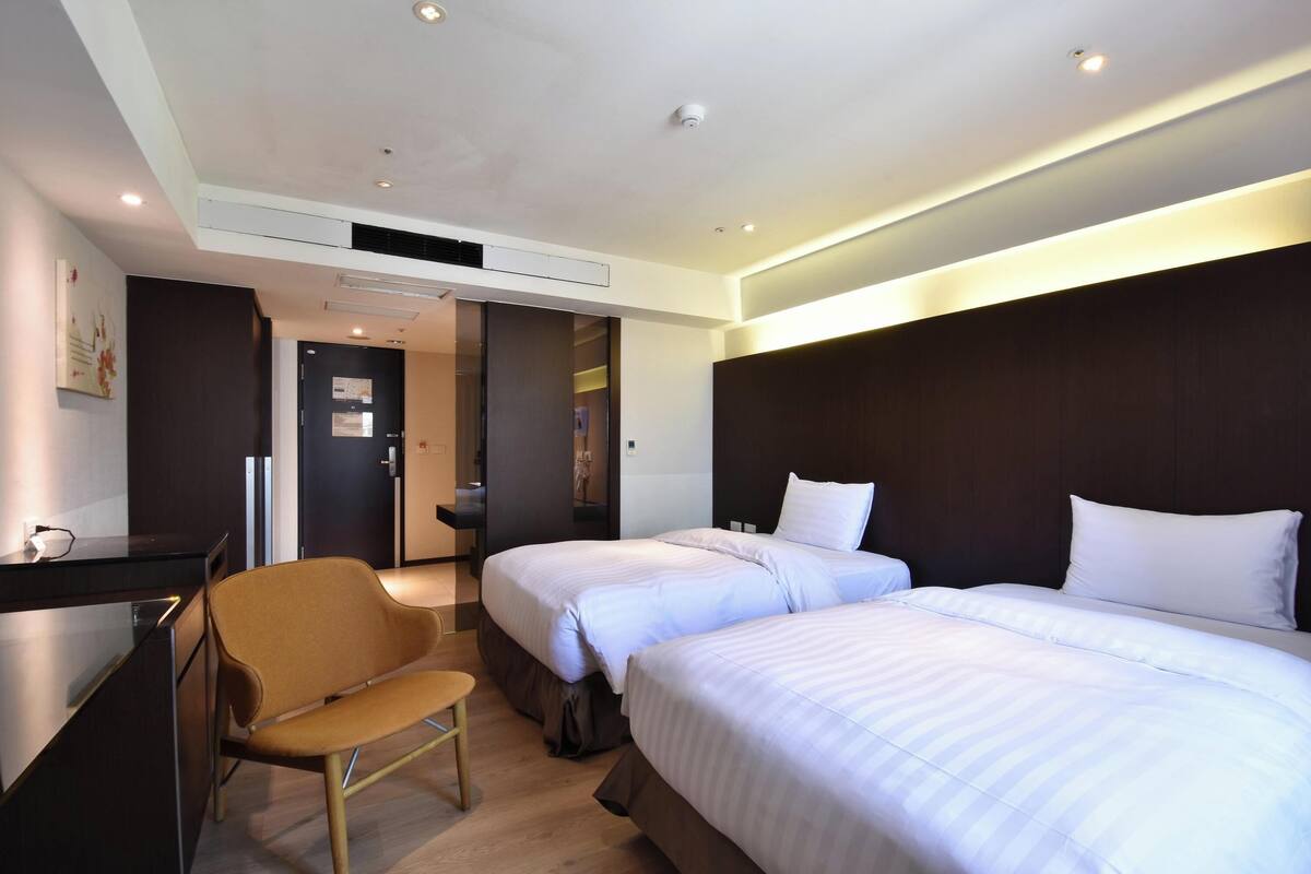 A hotel room with two beds, a desk, a chair, and a door leading to a bathroom.