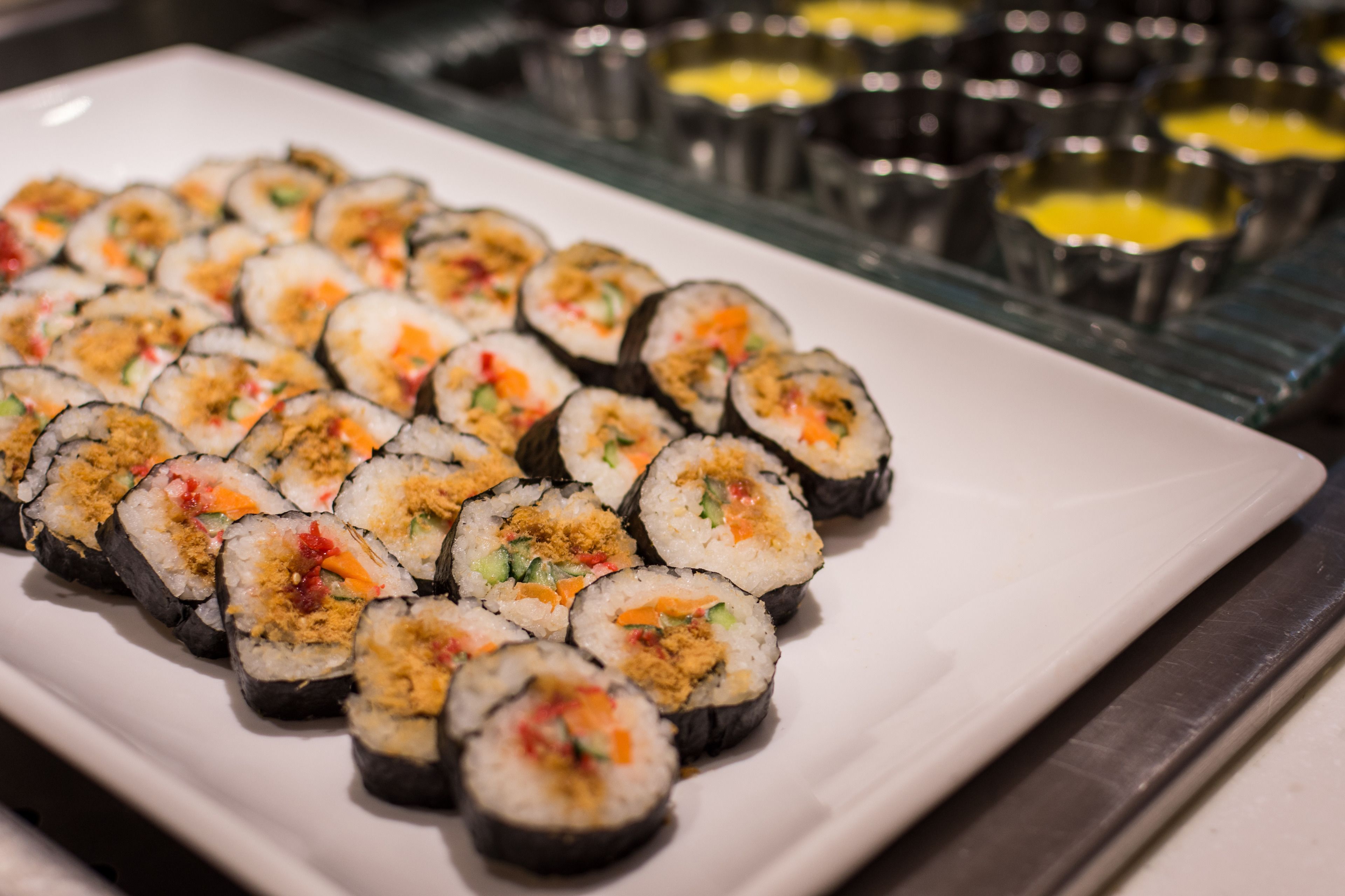 Um prato branco com sushi disposto com cuidado, acompanhado por pequenas tigelas de molhos de cobertura ao fundo.