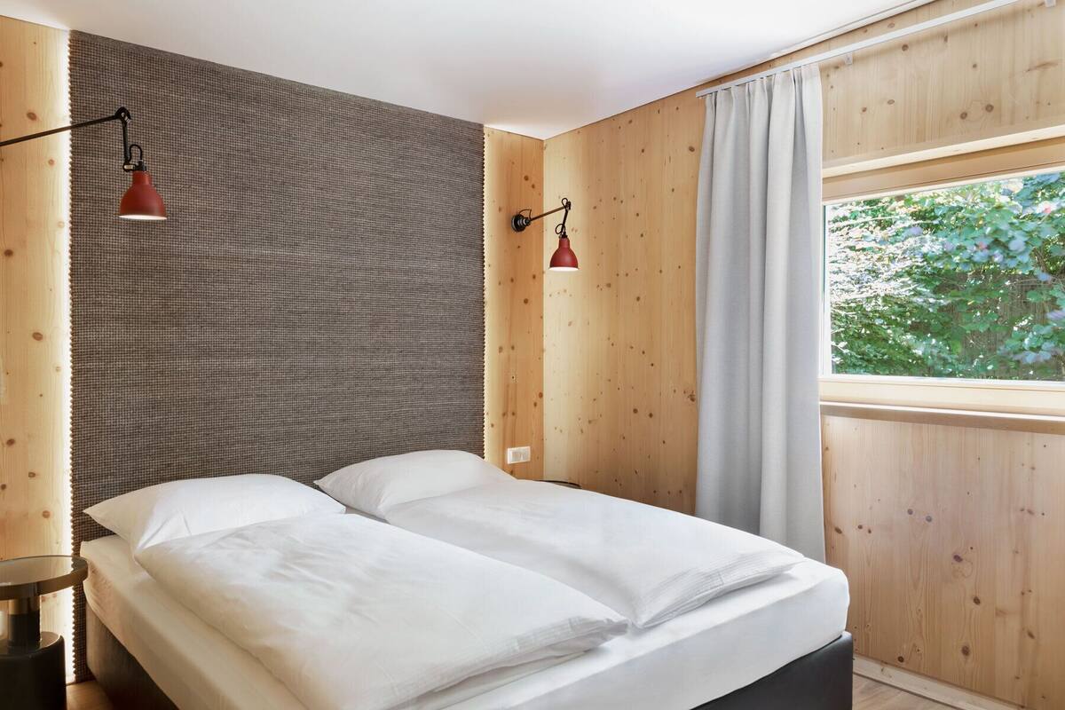 A bedroom with a bed, two wall-mounted lamps, a window with curtains, and a view of greenery outside.