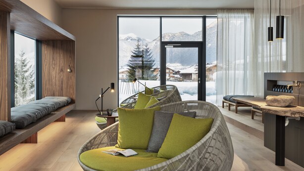 A modern living room with a large window, a wicker sofa, a wooden bench, and a small table with a book. - natürlich Hell. Aparthotel & Camping (Fuegen)