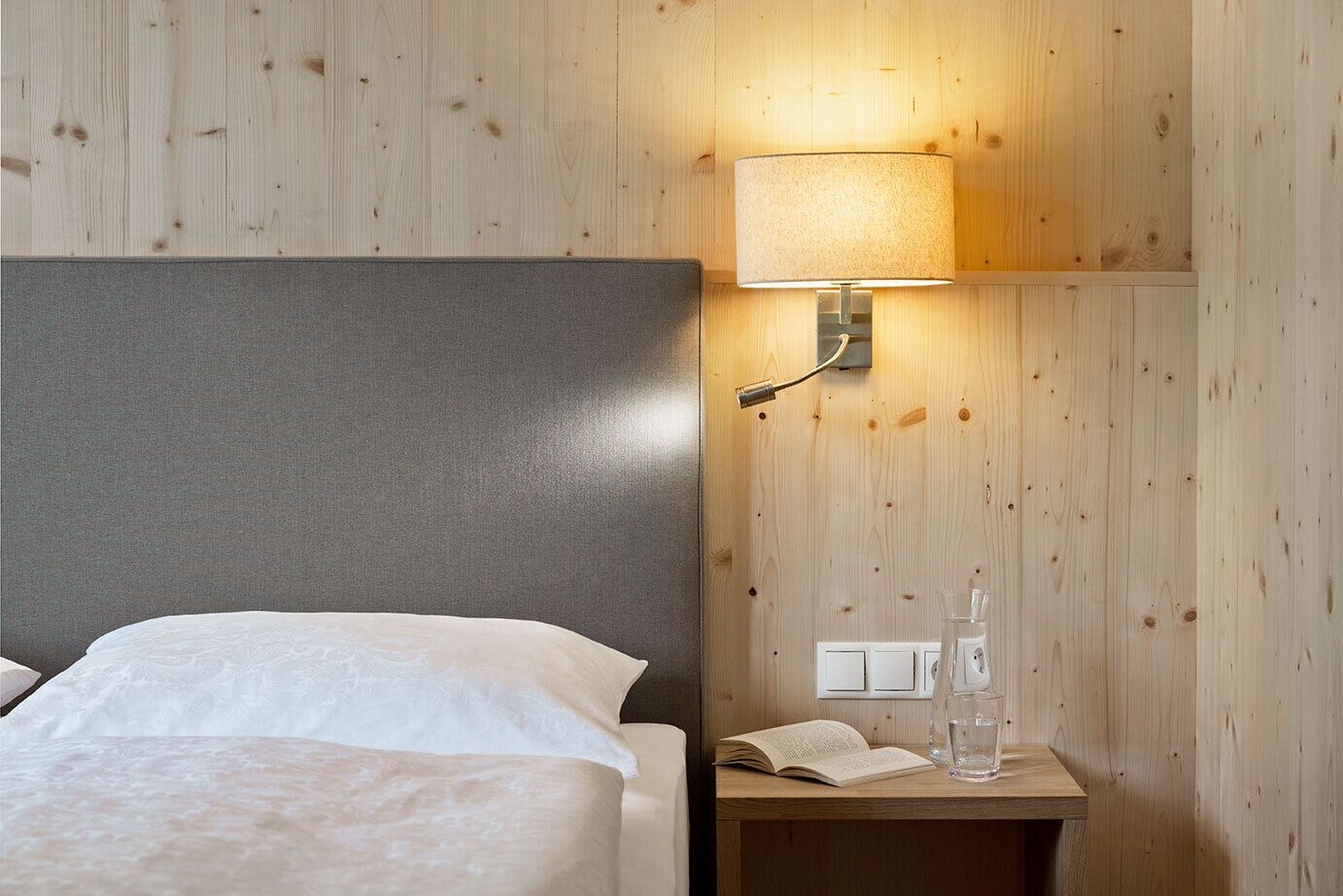 Habitación de hotel con pared de madera, cama con ropa blanca, mesita de noche con lámpara, un libro y un vaso de agua.