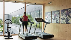 A gym with treadmills, a stationary bike, and a large window with a view of a building and trees. - natürlich Hell. Aparthotel & Camping (Fuegen)