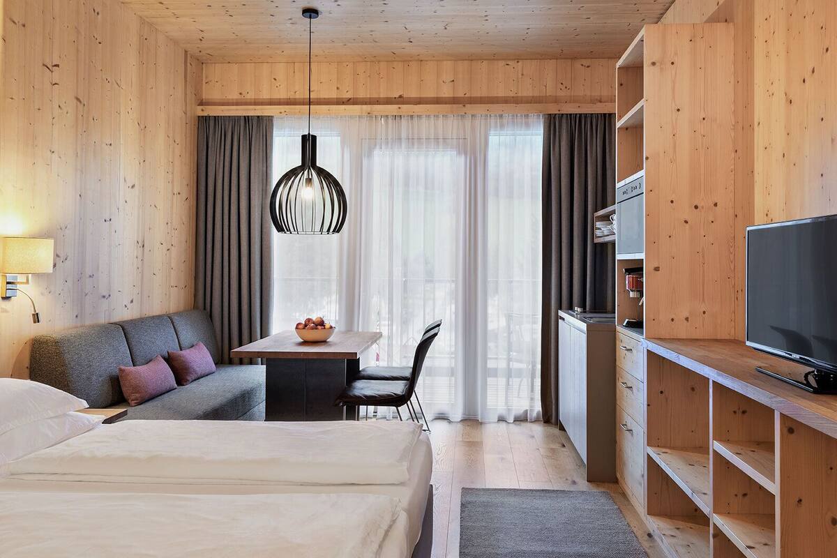 A hotel room with a bed, a sofa, a dining table with a bowl of fruit, a TV, and wooden walls.