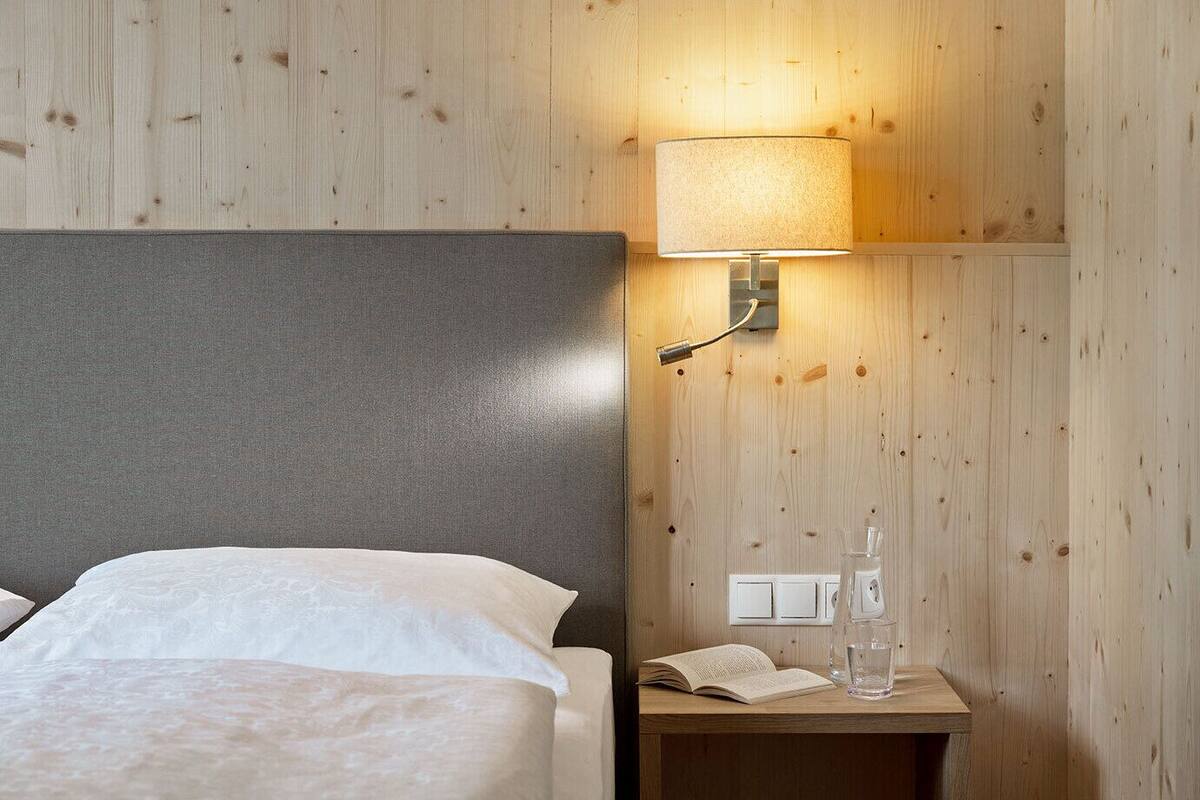 A hotel room with a wooden wall, a bed with white linens, a nightstand with a lamp, a book, and a glass of water.
