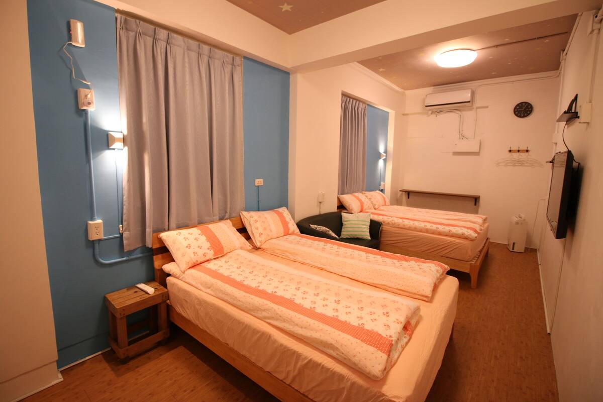 A bedroom with two beds, a nightstand, a small table, and a wall-mounted TV.