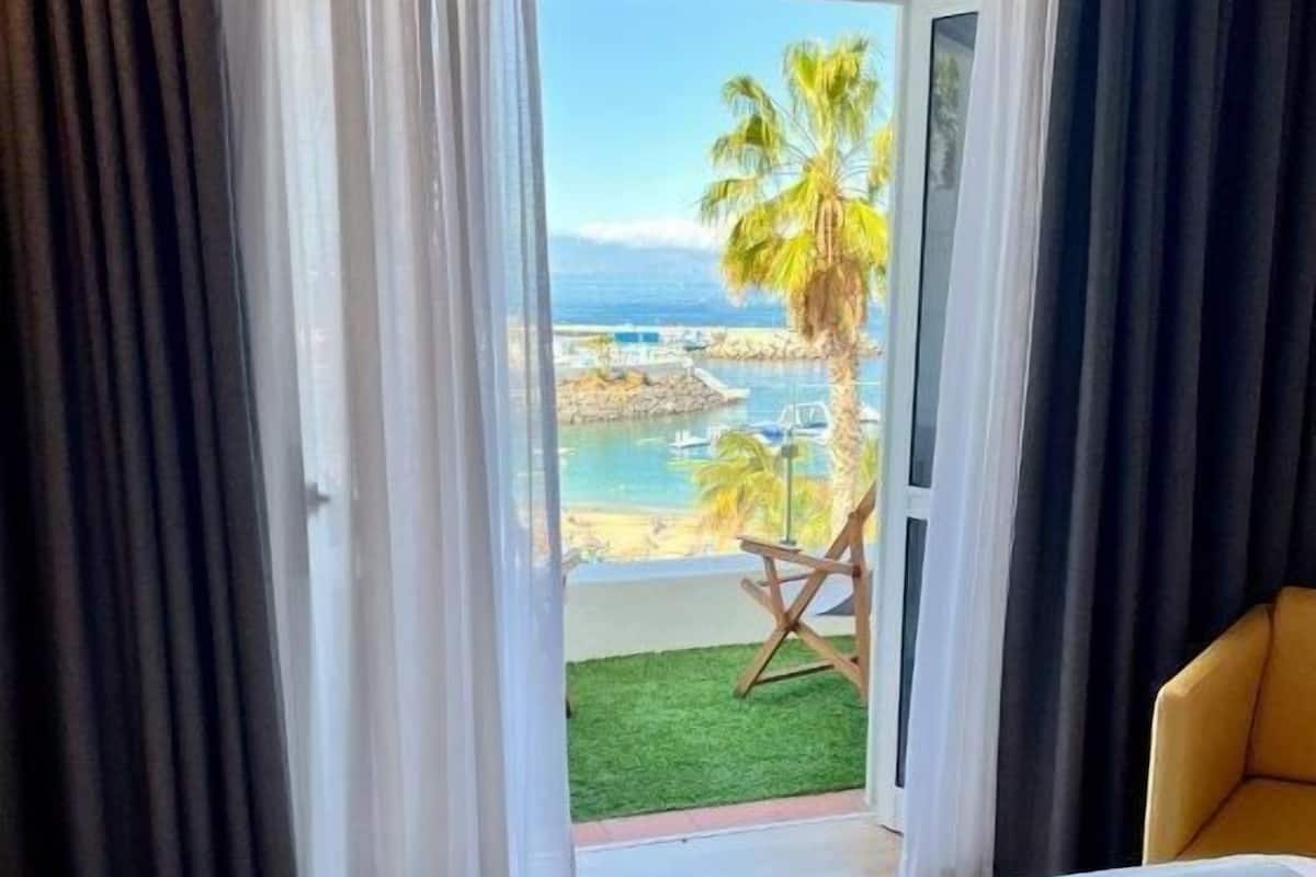 A hotel room with a bed, a chair, and a view of a beach and palm trees.