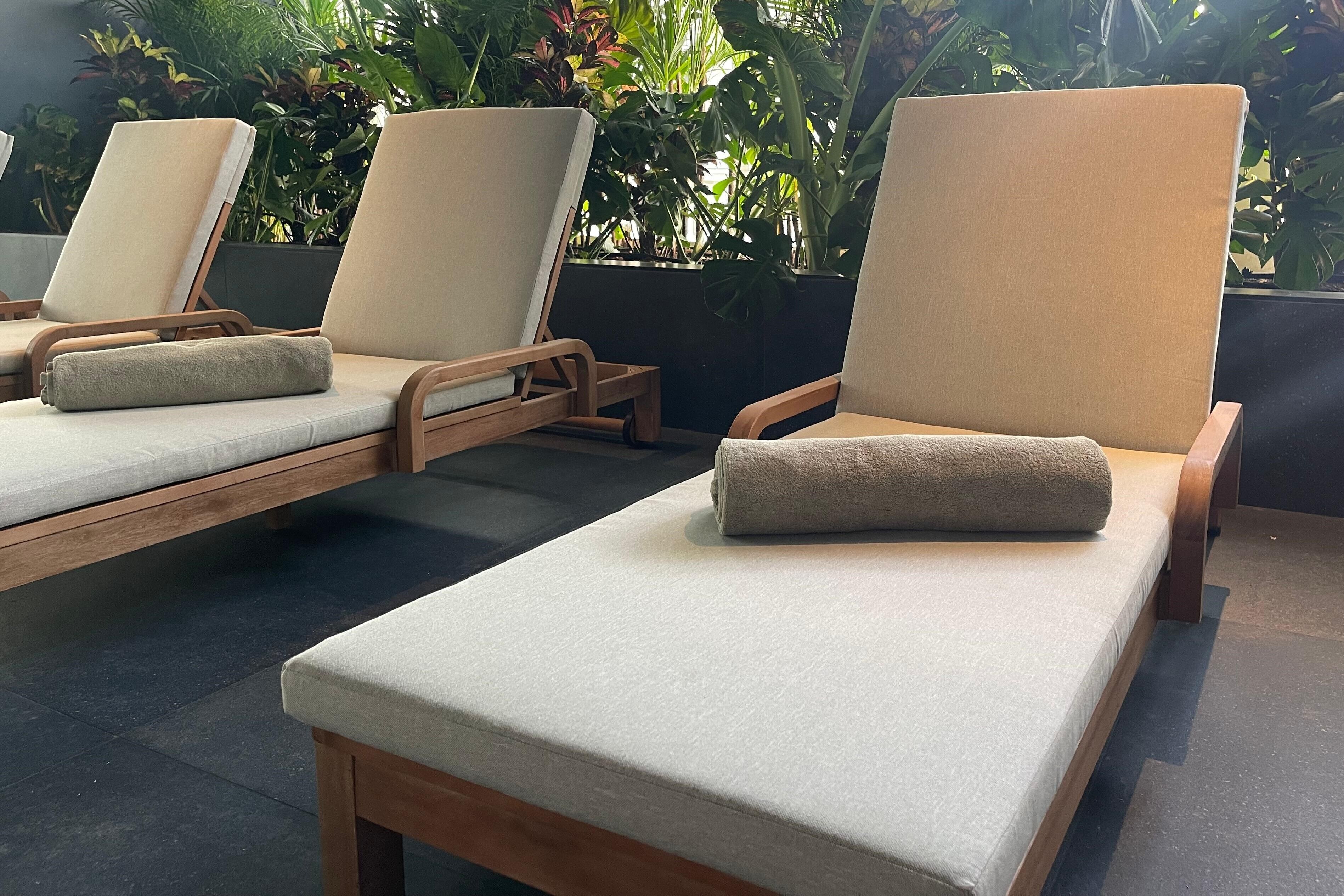 A row of wooden lounge chairs with beige cushions, arranged on a tiled floor with a backdrop of lush green plants.