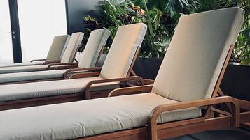 A row of wooden lounge chairs with beige cushions, positioned in a sunlit area with large windows and lush green plants in the background.