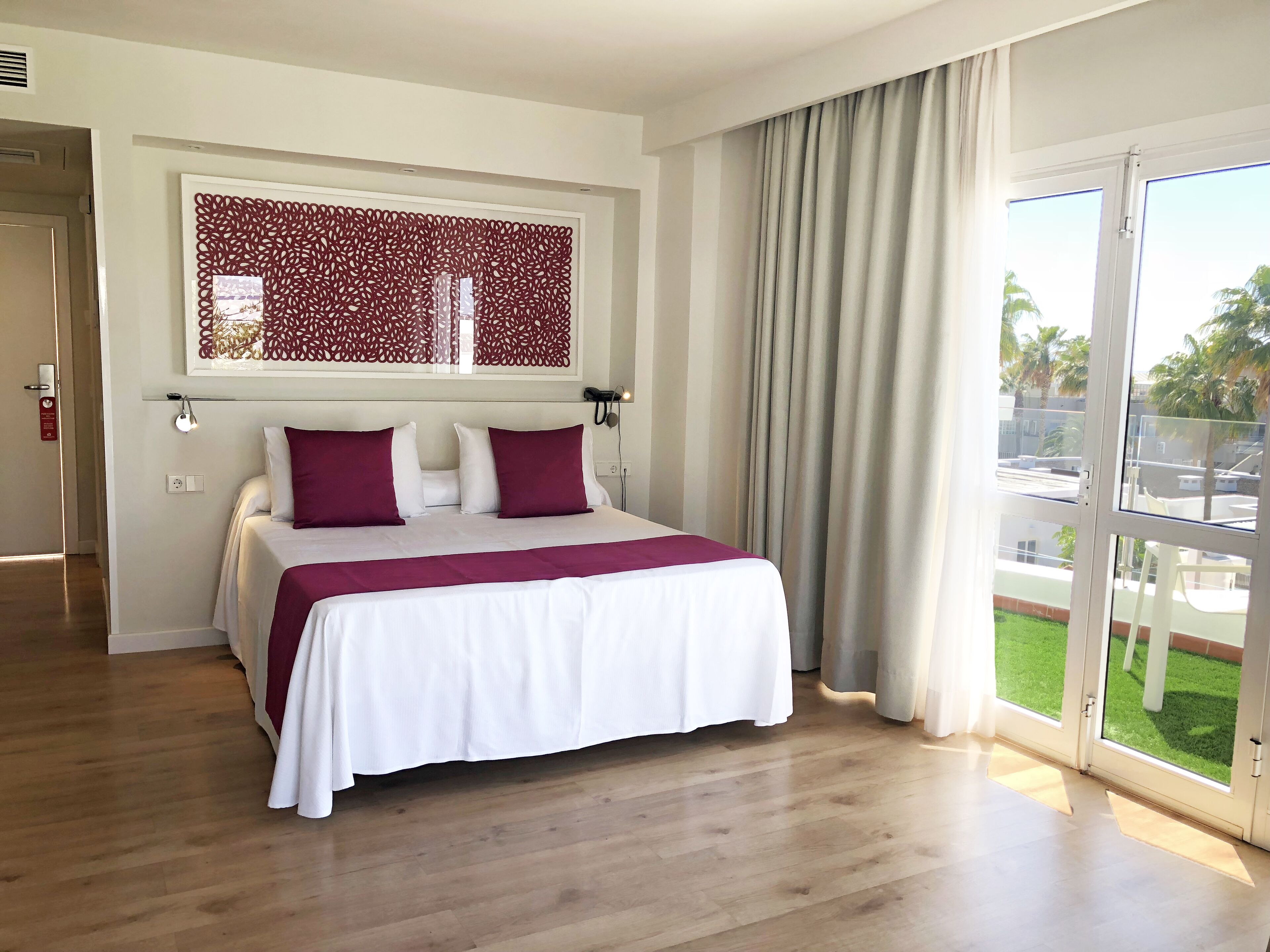 Ett hotellrum med en stor säng, en dekorativ v äggpanel, utsikt över omgivningarna genom en skjutdörr och ett trägolv.