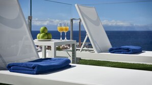 Un área de descanso al aire libre iluminada por el sol, con una mesa dispuesta con manzanas verdes y vasos de jugo de naranja, con vista al mar.