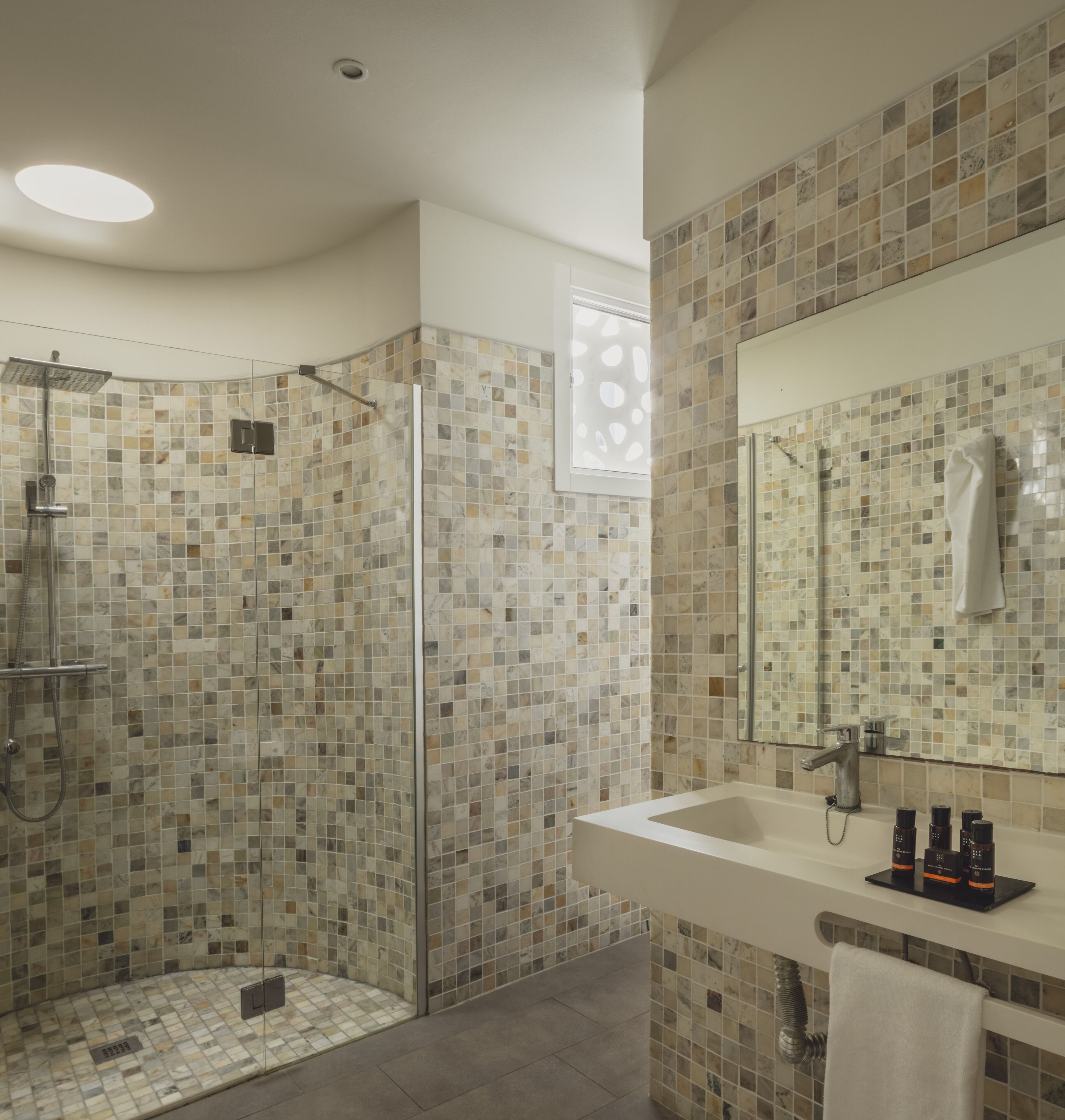 Un baño moderno con ducha envuelta en cristal, un espejo grande y un lavamanos blanco con artículos de aseo.