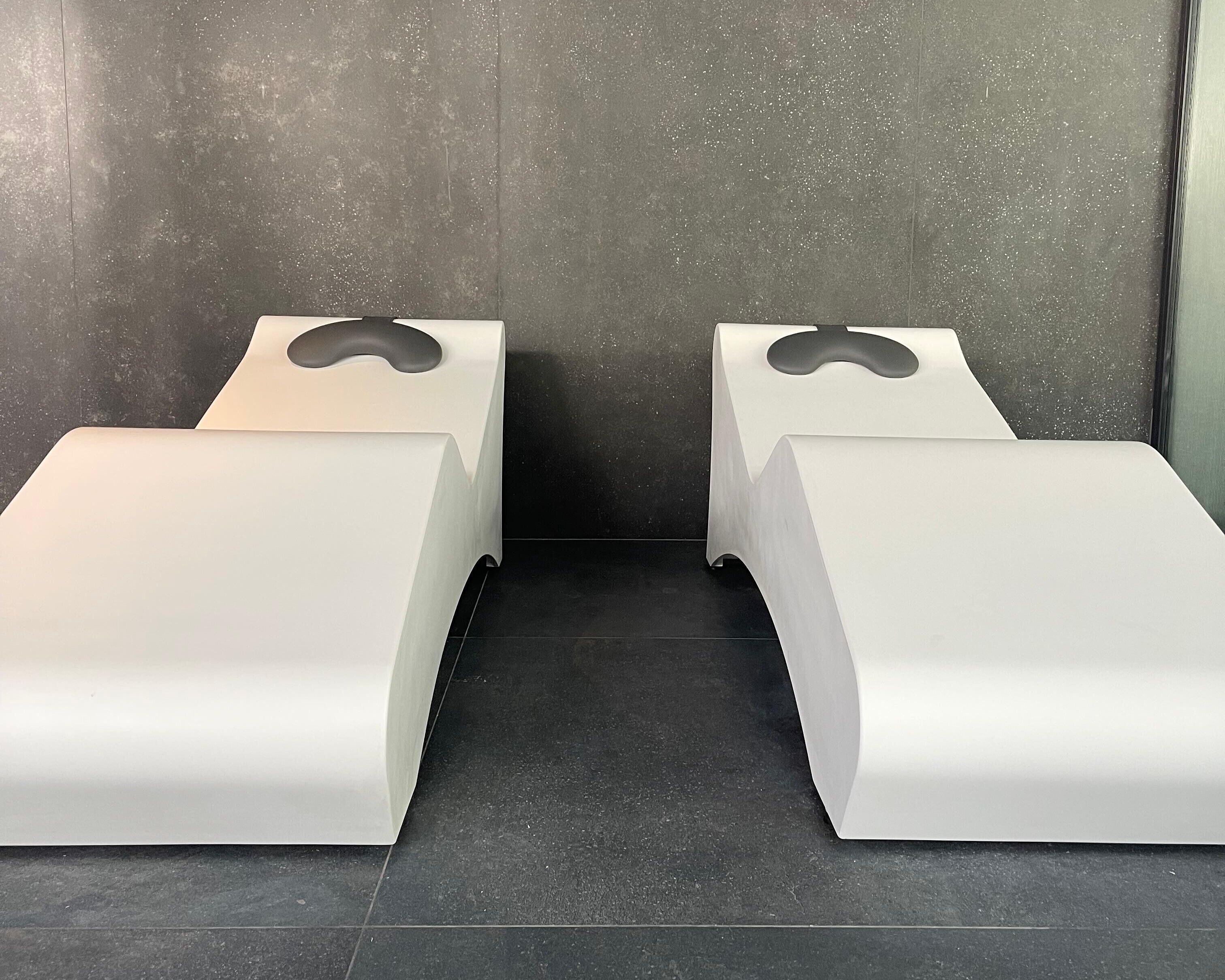 Zwei weiße, moderne Sitzbereiche im Bench-Stil mit dunklem Fliesenboden und einer schlichten Wand im Hintergrund.
