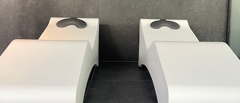 Zwei weiße, moderne Sitzbereiche im Bench-Stil mit dunklem Fliesenboden und einer schlichten Wand im Hintergrund.