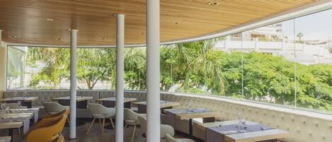 Un restaurante moderno con techos altos, vigas de madera y amplios ventanales que ofrecen vistas a zonas verdes y edificios.