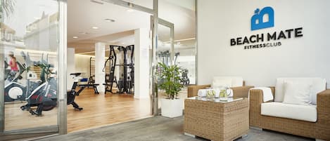 Interior de un gimnasio con equipo de ejercicio, una zona de recepción y un área de descanso con una planta.