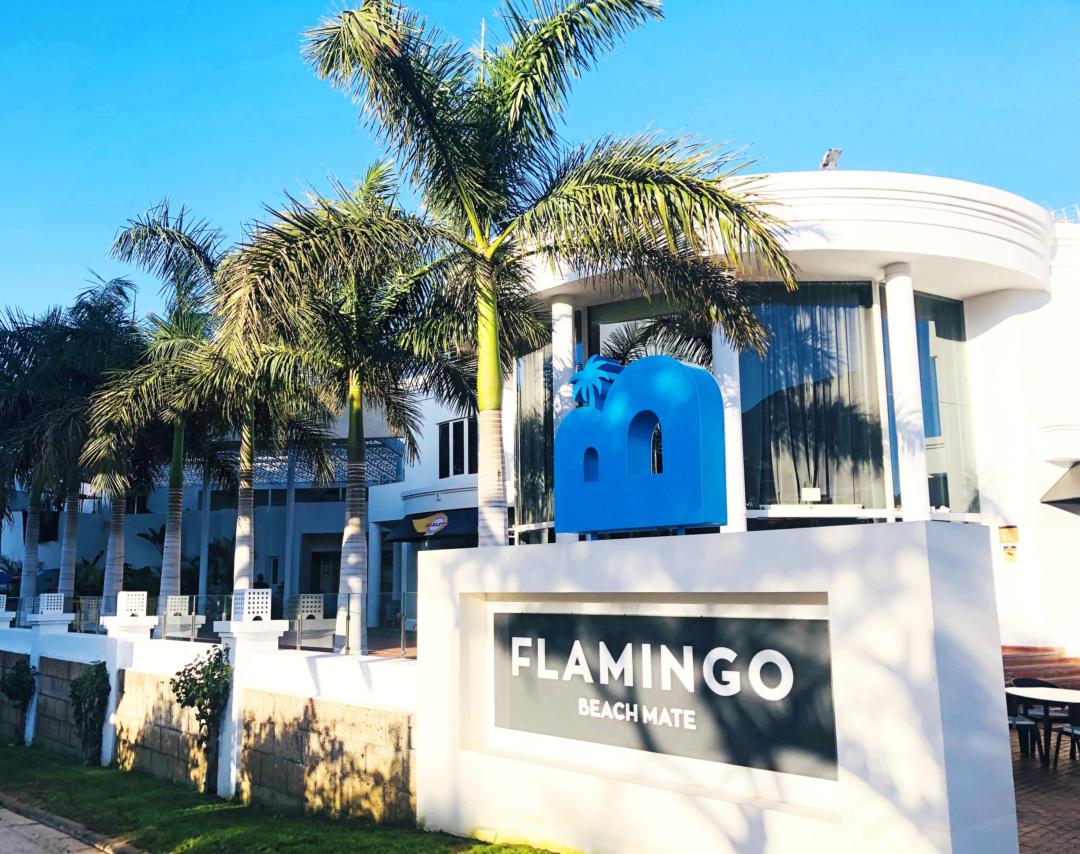 Un letrero que dice 'FLAMINGO BEACH MATE' con palmeras y un edificio moderno al fondo.