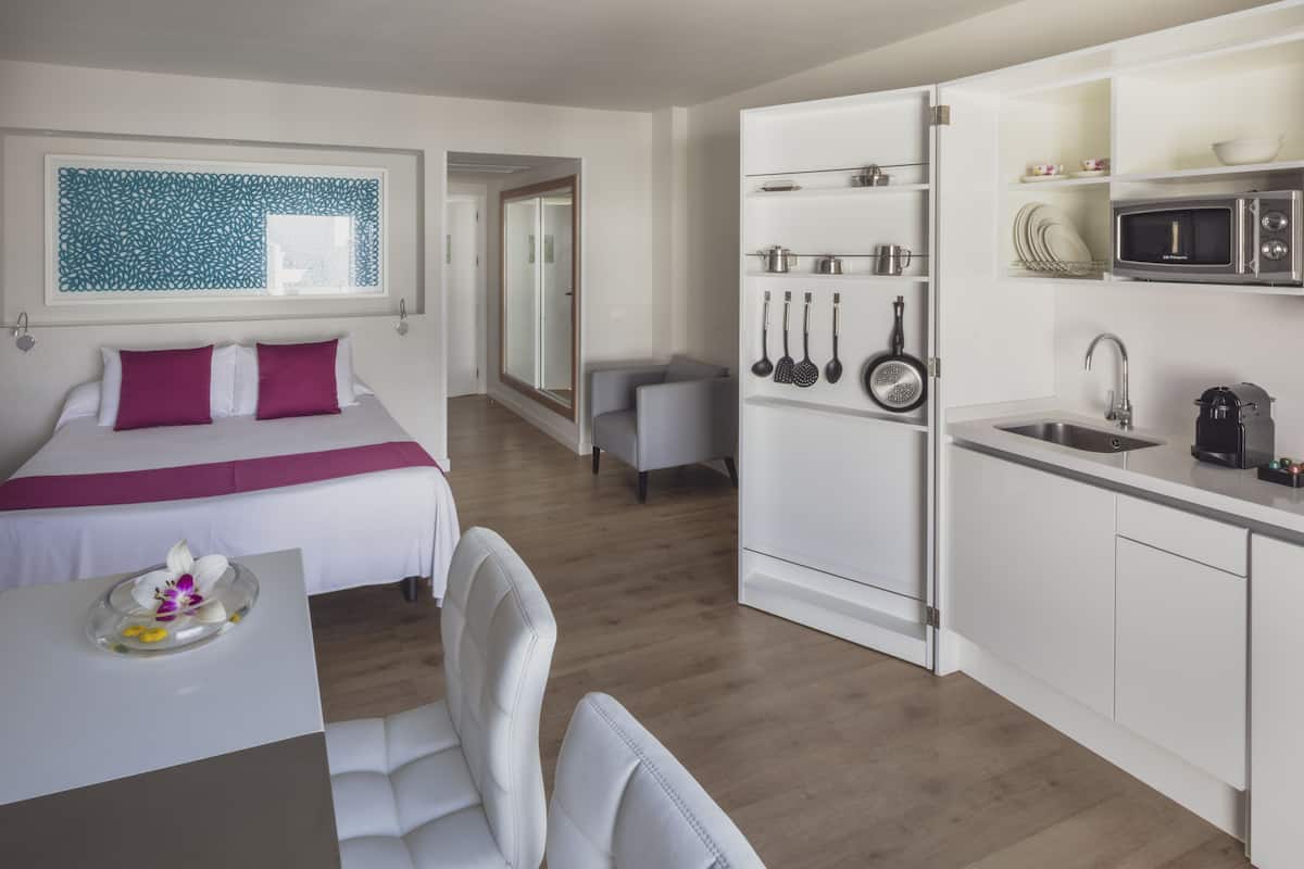 A modern hotel room with a bed, a desk, a chair, a kitchen area with a microwave, sink, and utensils.