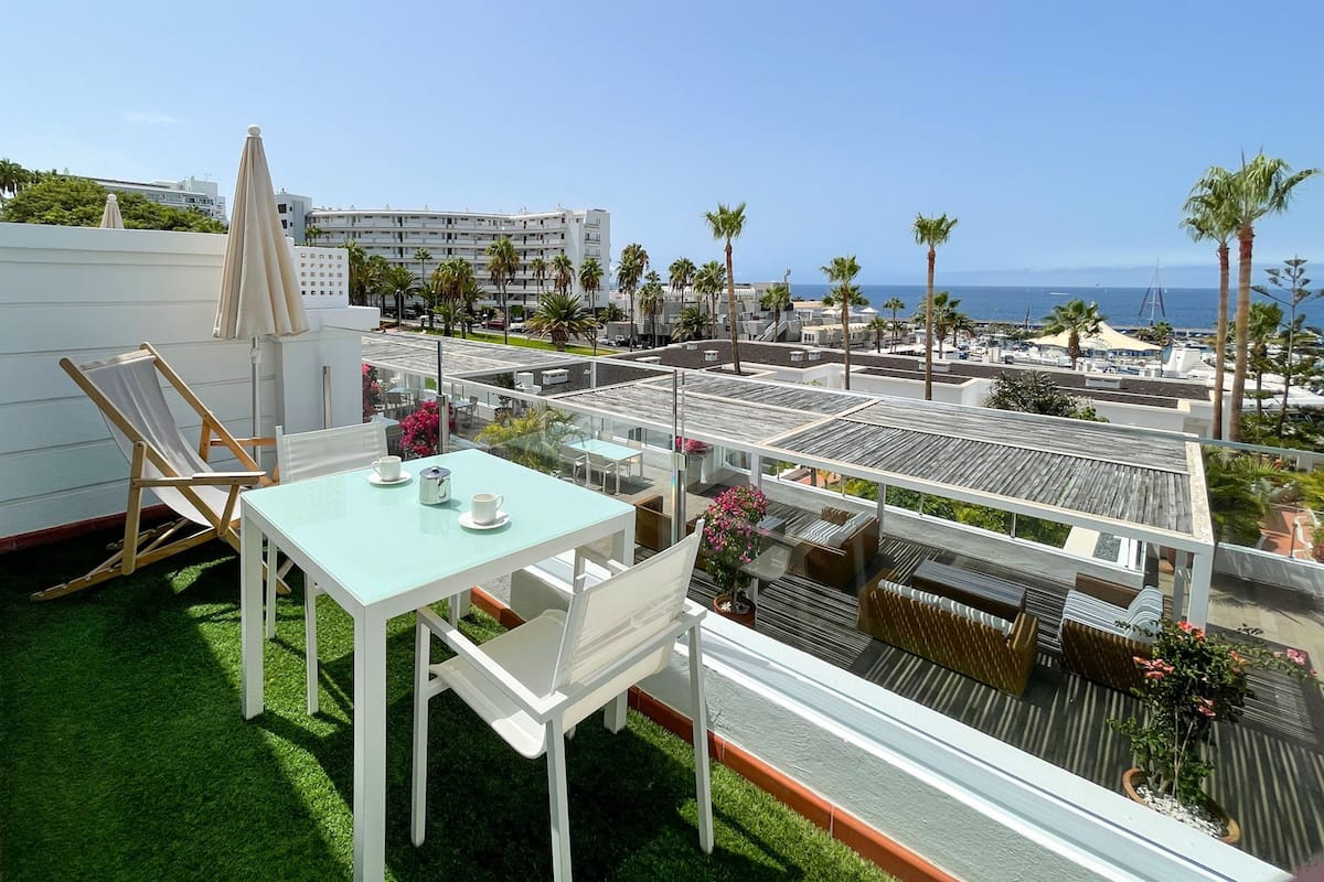A rooftop terrace with a table and chairs, a lounge chair, and a view of the ocean and buildings.