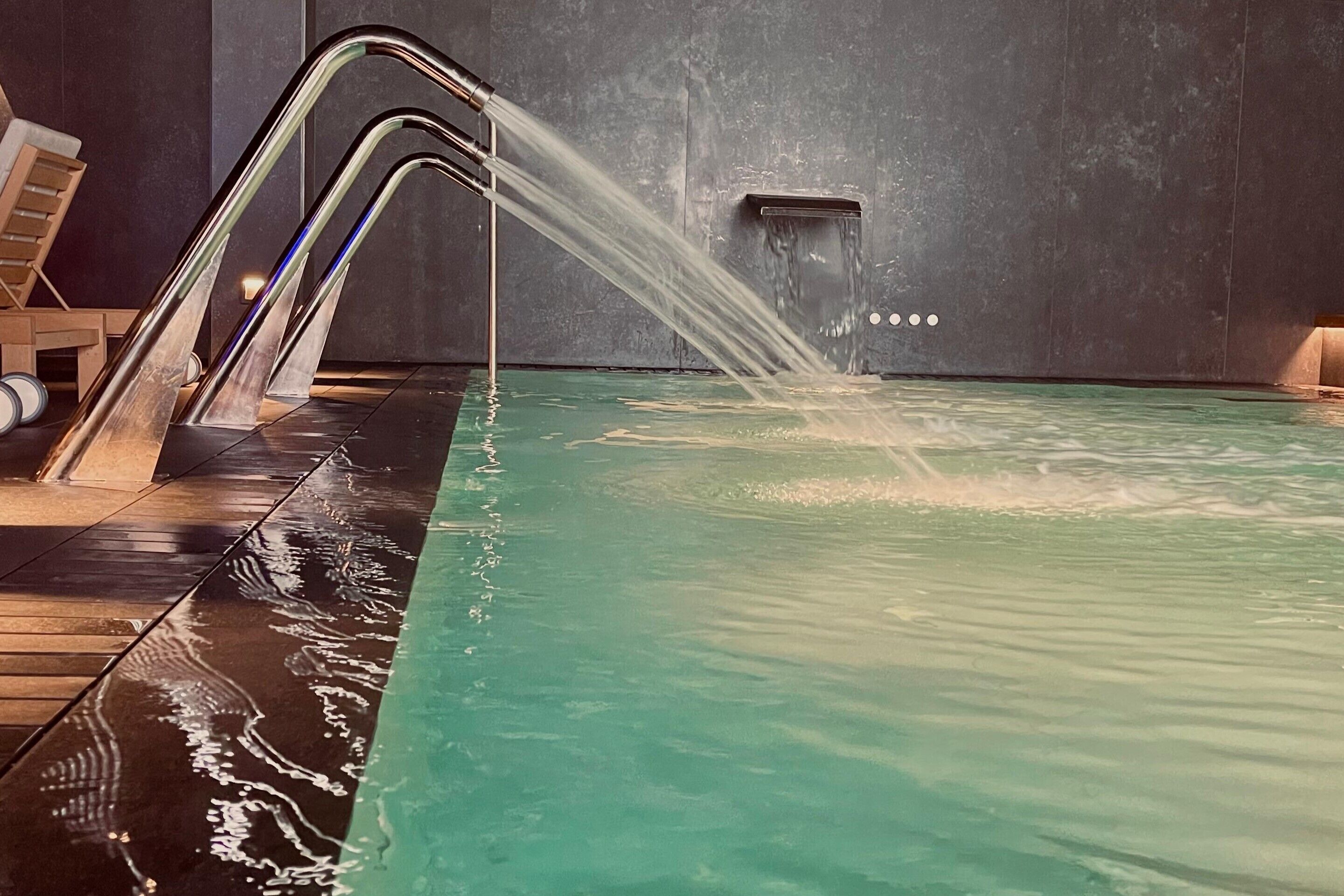 A pool with water flowing from a modern faucet, a wooden lounge chair, and a towel rack.