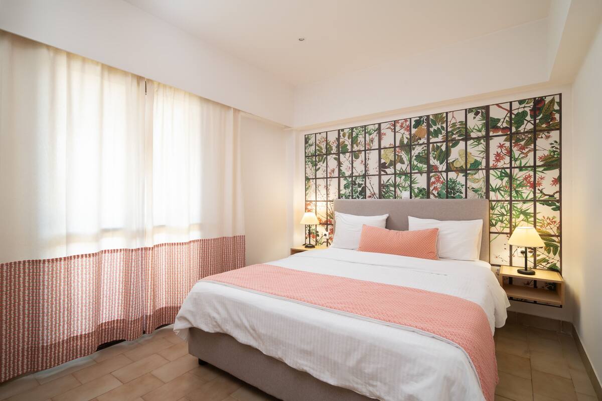 A hotel room with a large bed, two bedside lamps, a window with sheer curtains, and a decorative wall panel.