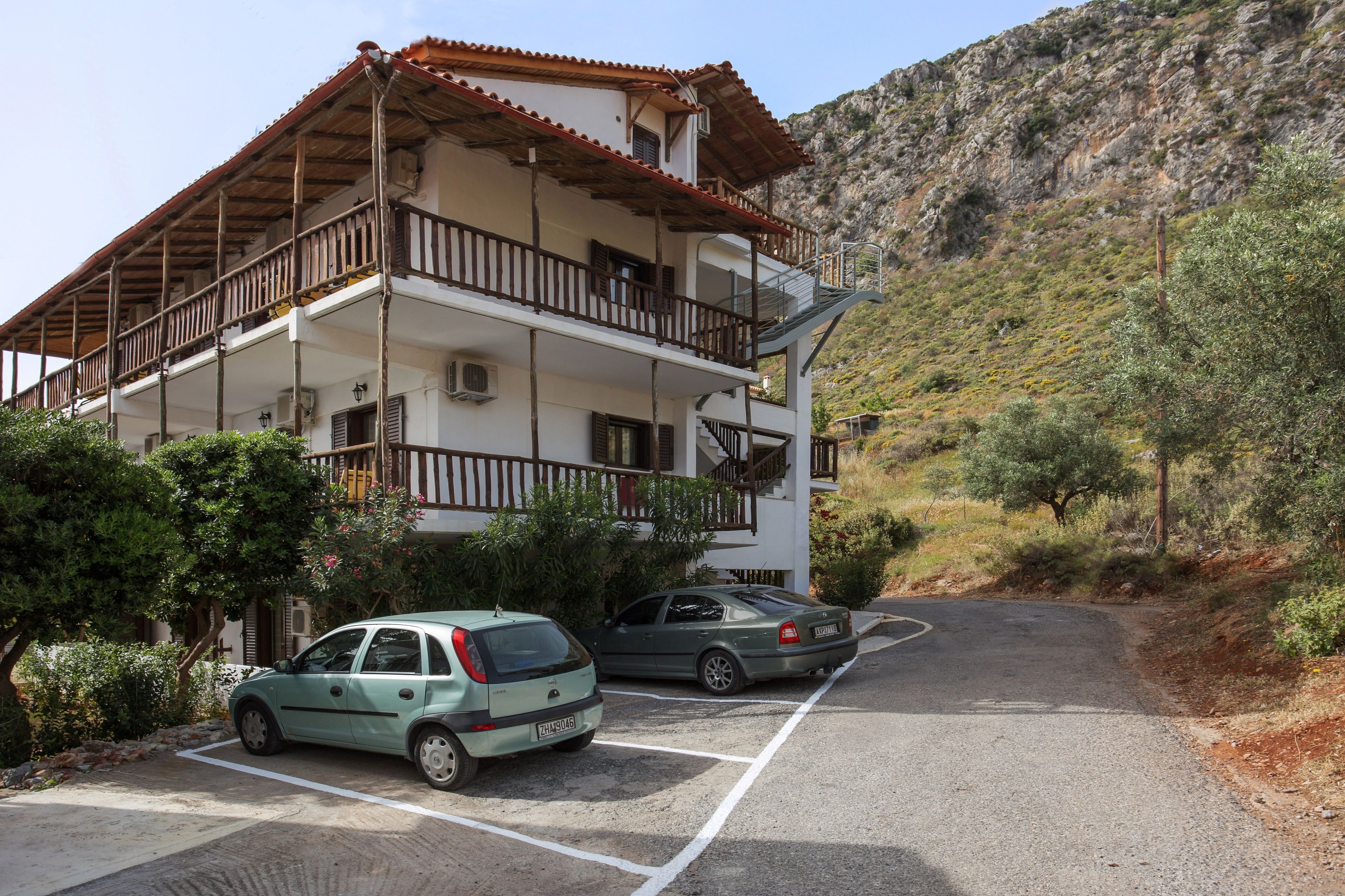 A multi-story building with balconies, parked cars, and a mountainous backdrop.