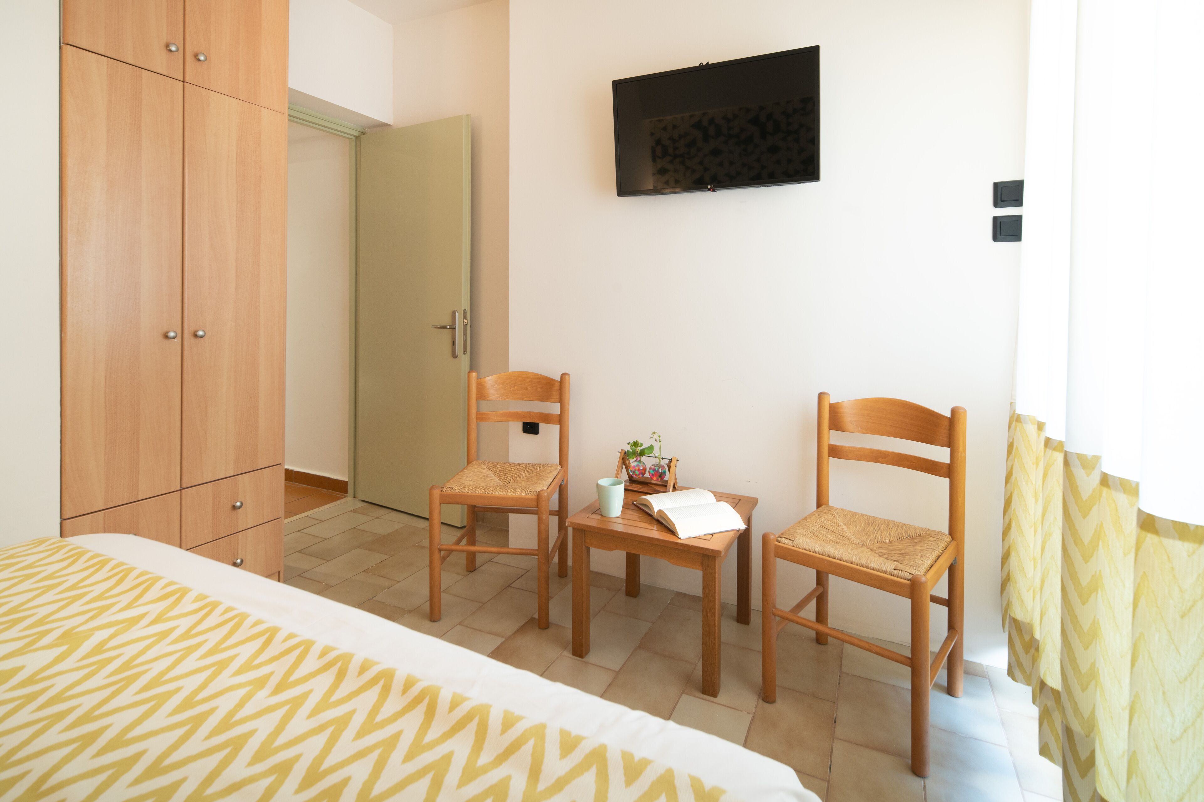 Ein Schlafzimmer mit einem Bett, zwei Holzstühlen, einem kleinen Tisch mit Buch und Tasse, einem an der Wand befestigten Fernseher und einem Kleiderschrank.