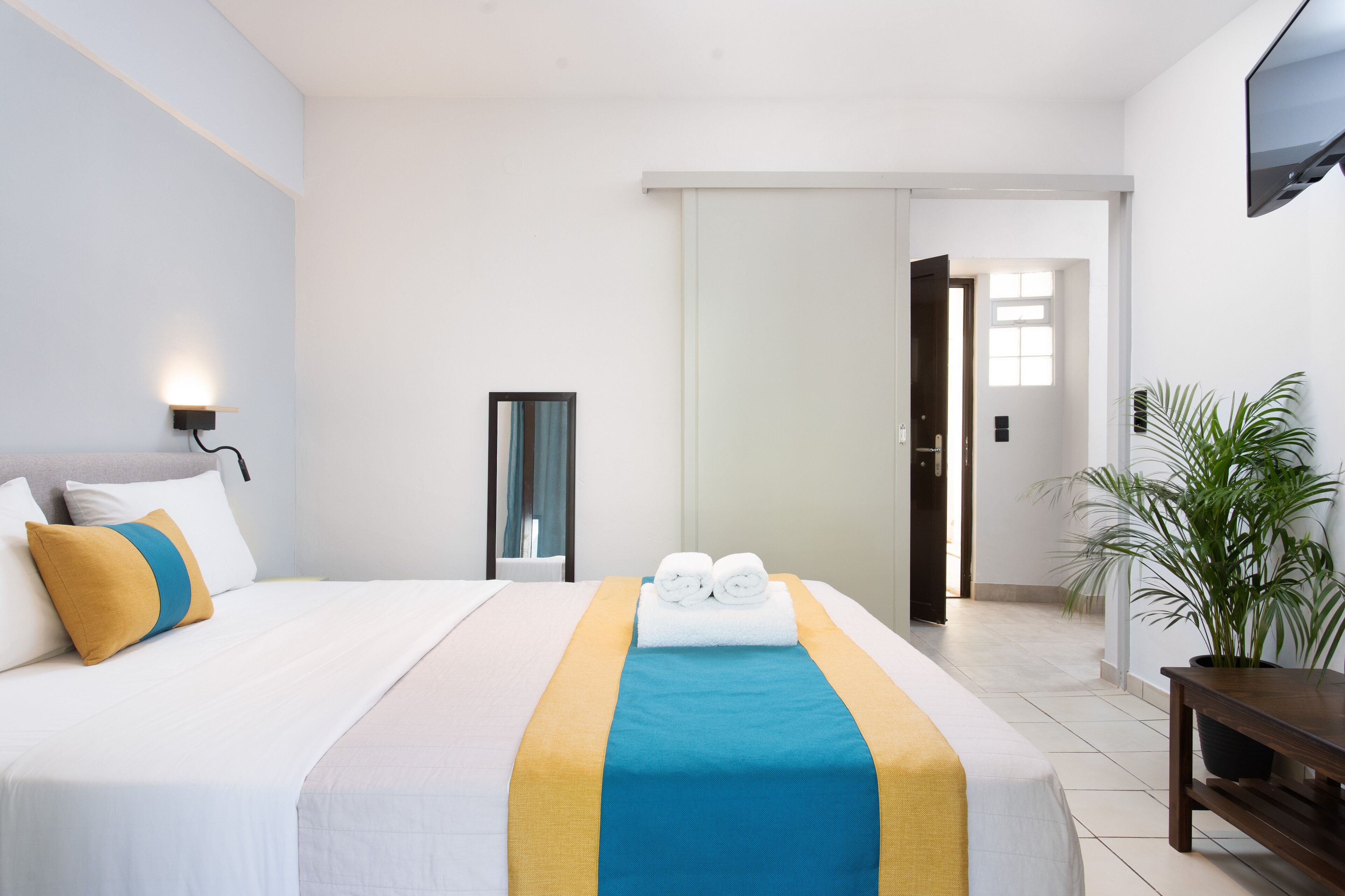 Habitación de hotel con una cama bien hecha, una mesita de noche, una planta en maceta y un televisor en la pared.