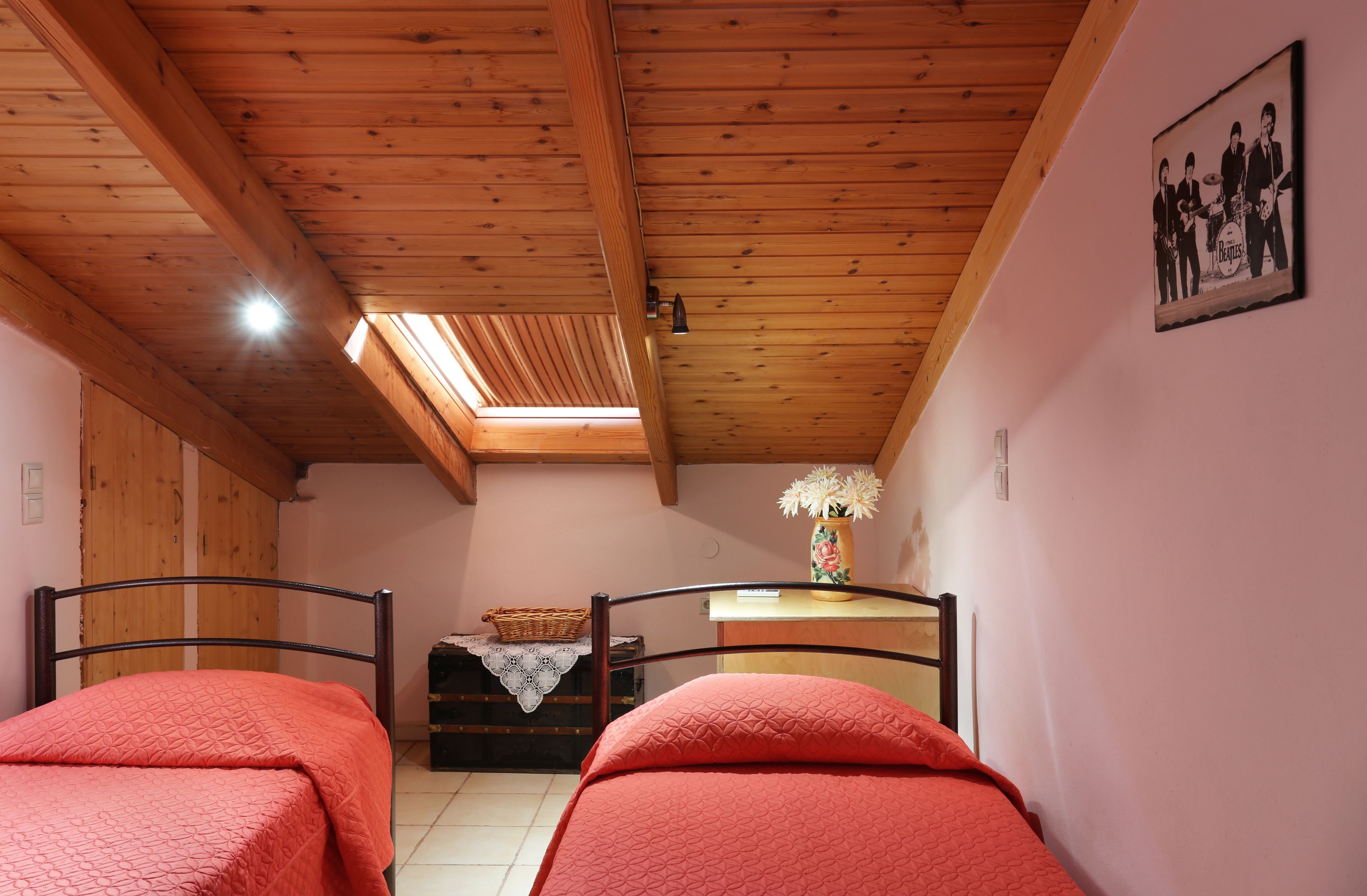 A bedroom with two beds, wooden ceiling beams, a skylight, a wall-mounted picture, and a small table with a vase.